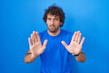 Hispanic young man standing over blue background moving away hands palms showing refusal and denial...