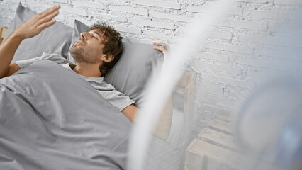 Hispanic man with beard enjoys breeze from fan while lying in bedroom, eyes closed in relaxation