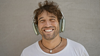 A cheerful young hispanic man with curly hair, wearing headphones and a necklace, poses against a plain white background.