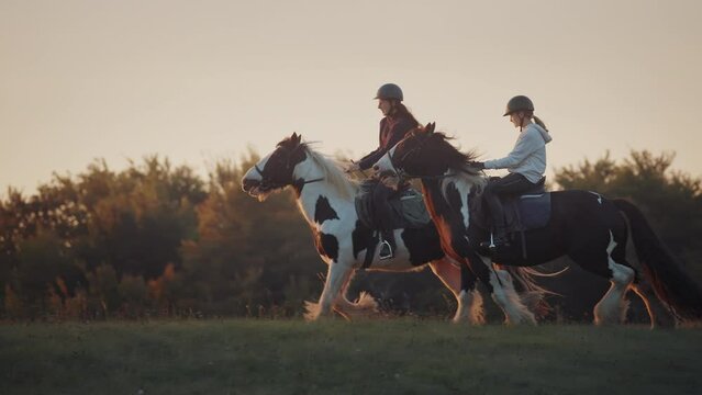 Two young women horseback riding in the green countryside at golden sunset. Riding on a horse in the field. Slowmotion shot.