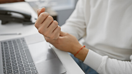 A young adult man with a beard experiences wrist pain while using a laptop in an office setting.