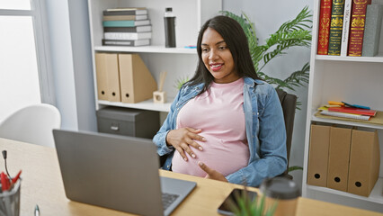 Expecting young business woman, smiling and touching belly while enjoying a successful video call at the office