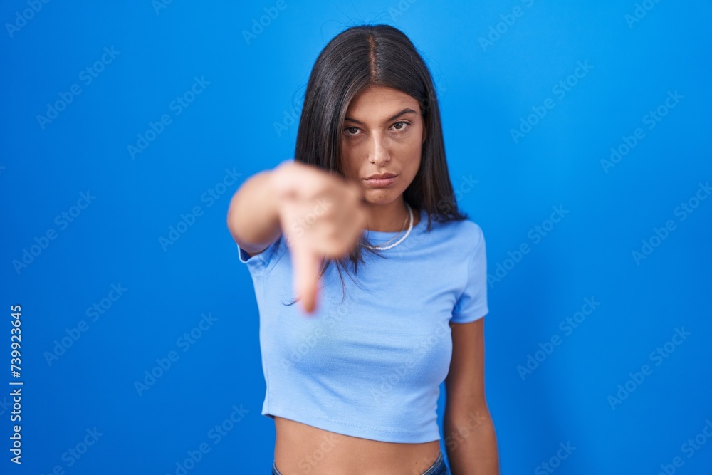 Poster Brunette young woman standing over blue background looking unhappy and angry showing rejection and negative with thumbs down gesture. bad expression.