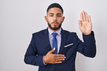 Young hispanic man wearing business suit and tie swearing with hand on chest and open palm, making a loyalty promise oath