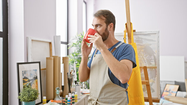 Bearded man drinking coffee in a bright art studio filled with paintings and art supplies.