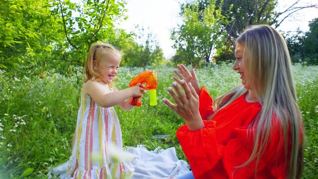 A woman and a little girl are happily playing with soap bubbles in a field of flowers on a sunny summer day, surrounded by nature and tall grass