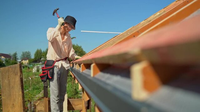 A worker hammers nails into boards. Roof construction. Roof sheathing. A young roofer in a shirt, a hat and glasses.
