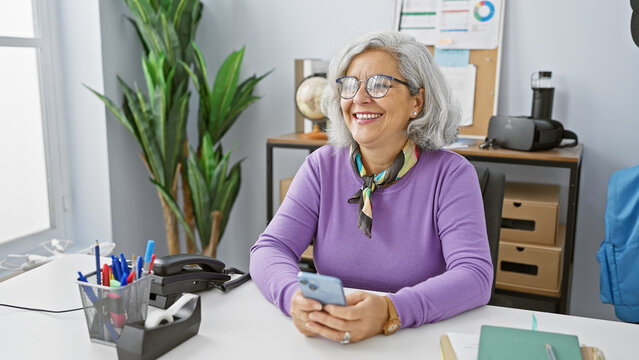 Smiling mature woman with glasses holding smartphone in modern office setting, exemplifying active elderly professionals.