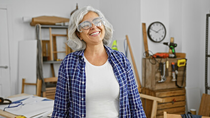 A cheerful middle-aged woman with grey hair wearing safety glasses in a carpentry workshop.