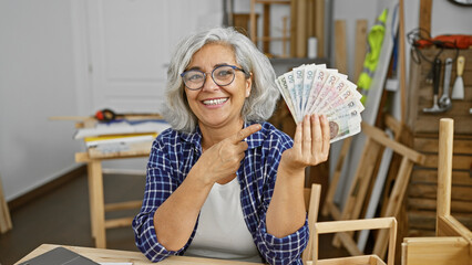 Mature woman holding polish zloty in a carpentry workshop, radiating joy and success.