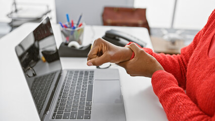 African woman in red experiences wrist pain while working with a laptop in a modern office setting.