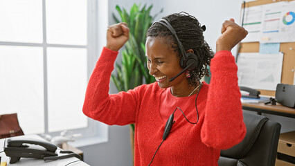 An african american woman celebrates with joy in her office, wearing a headset and a red sweater.