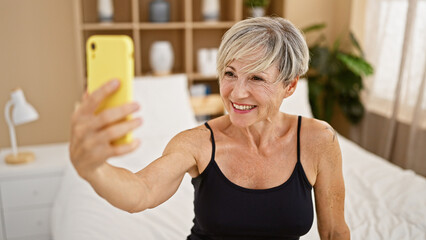 A cheerful mature woman takes a selfie in a bright bedroom, embodying positivity and modern...