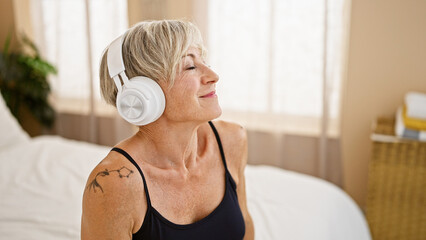 A relaxed mature woman with grey hair enjoying music on headphones in a cozy bedroom setting.