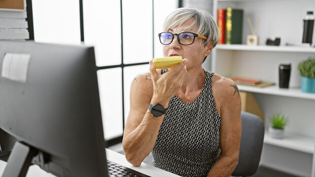 A mature woman with grey hair speaks into a phone in a modern office environment, depicting a professional setting.