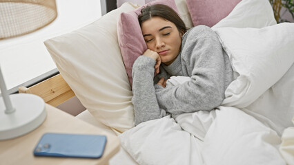 A young hispanic woman sleeping peacefully in a cozy bedroom setting with a smartphone nearby