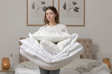 Woman holding pile of soft white folded duvet and pillows at home in her bedroom, cozy domestic...