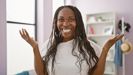 Cheerful african american woman with curly hair in a casual white shirt, laughing indoors at home with a bookshelf and a hat in the background. - Powered by Adobe
