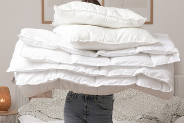 Woman holding pile of soft white folded duvet and pillows at home in her bedroom, cozy domestic...