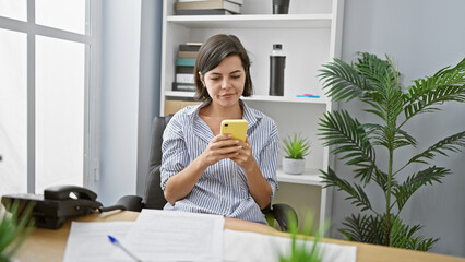 Successful young hispanic businesswoman, relaxed and concentrated, sitting at her office desk, working on her smartphone, a portrait of a beautiful woman with a short hairstyle.