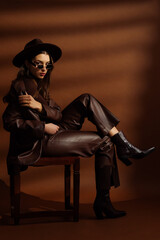Fashionable confident woman wearing hat, sunglasses, trendy  faux leather suit blazer, pants,  pointed toe ankle boots, posing on brown background. Full-length studio fashion portrait 