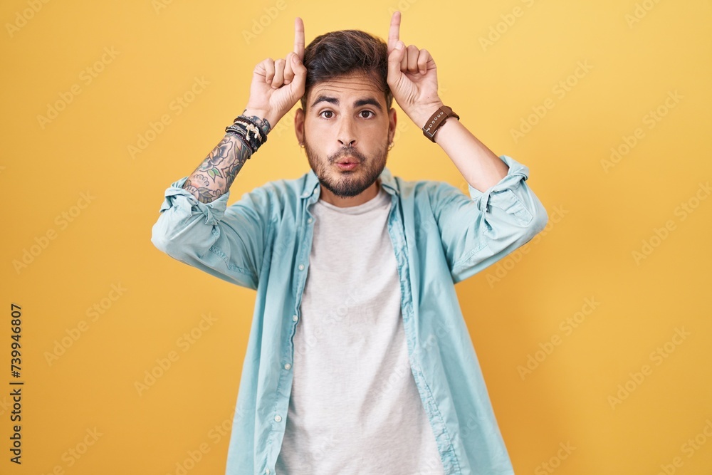 Sticker young hispanic man with tattoos standing over yellow background doing funny gesture with finger over