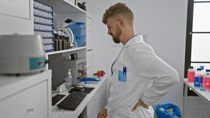 A young, bearded man with blue eyes wearing a lab coat grimaces with back pain in a clinical...