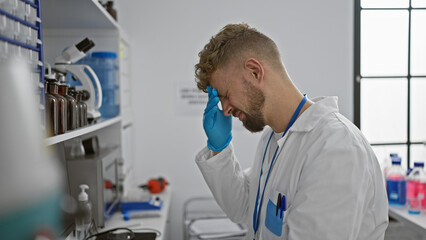 A stressed young man in a lab coat exhibiting a headache against a clinical laboratory backdrop