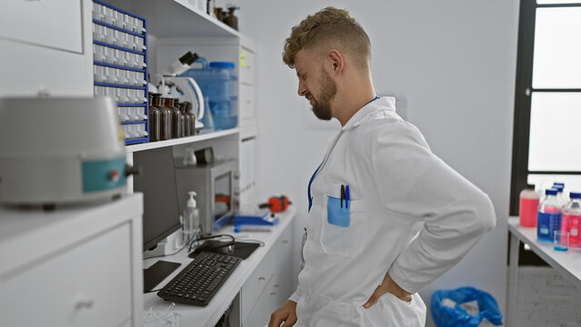 A young, bearded man with blue eyes wearing a lab coat grimaces with back pain in a clinical laboratory setting.