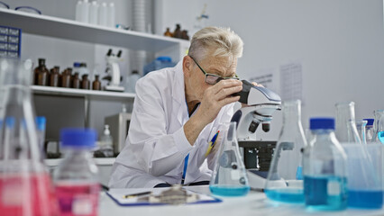 Dedicated grey-haired senior woman scientist immersed in intense microscope research work at phd...