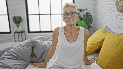 Serious-looking, grey-haired senior woman comfortably relaxing on her cozy bed, deep in thought, in...