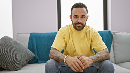 Fototapeta premium A confident young hispanic man with a beard sits casually in a modern indoor living room setting.