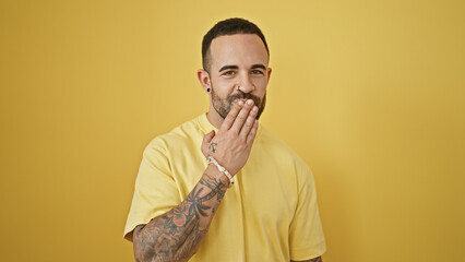 Handsome bearded man covering mouth with hand against a yellow background, expressing surprise or...
