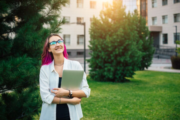 Female college student happily smiles holding laptop after classes on campus outdoors. Stylish young woman standing outside among green trees.