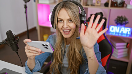 Smiling caucasian woman waving in a gaming room with headphones, microphone, and 'on air' sign.