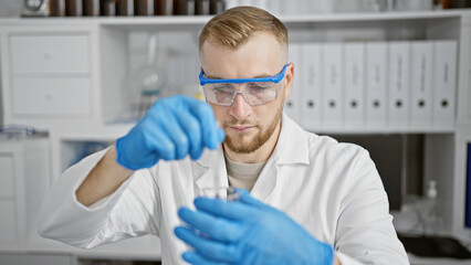 A focused caucasian man in a lab coat examines a test tube in a clinical laboratory setting, wearing protective eyewear and gloves.