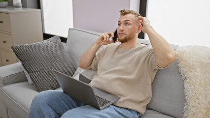 Handsome caucasian man multitasking with laptop and phone in modern apartment living room