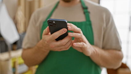 A young bearded man wearing an apron using a smartphone indoors in a blurred room.