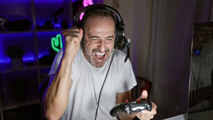 Middle age grey-haired male streamer joyfully celebrates his game win in a dim gaming room,...