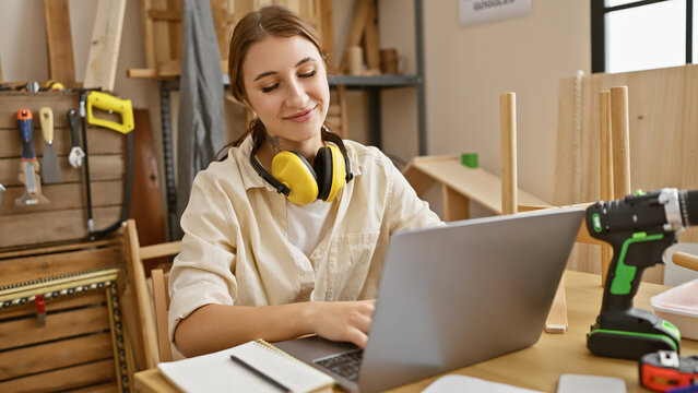 Brunette woman working on laptop in a carpentry workshop with headphones and tools around.