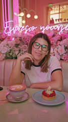 Smiling woman wearing glasses at a pink-themed cafe with neon sign, dessert and coffee on table, and floral backdrop