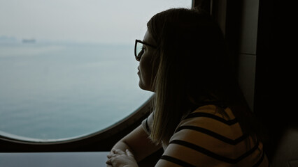 Silhouetted young woman wearing glasses looking out a large porthole on a sea journey, evoking...