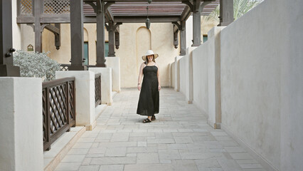 A smiling woman in a wide-brimmed hat and black dress strolls through the traditional architecture...