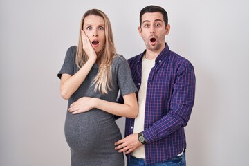 Young couple expecting a baby standing over white background afraid and shocked, surprise and amazed expression with hands on face