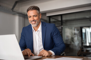 Smiling 40s mature Latin business man ceo trader using computer, typing, working in modern office,...