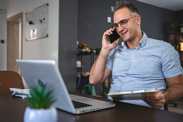 Middle aged Caucasian man talking on the phone while working on laptop at home