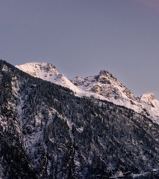 Winter magic in the Swiss Alps: A village, snowy forest, and pink-hued peaks under the evening sun. A grand glacier adds to the picturesque panorama.