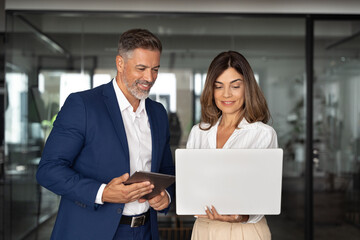 Fototapeta na wymiar Mature 40s Latin business man and European business woman discussing project on laptop gadget in office. Two diverse partners, colleagues, team of confident professional business people work together.