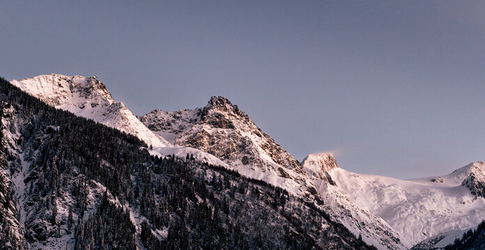 Winter magic in the Swiss Alps: A village, snowy forest, and pink-hued peaks under the evening sun. A grand glacier adds to the picturesque panorama.