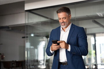Smiling mature Latin or Indian businessman holding smartphone in office. Middle aged manager using cell phone mobile app. Digital technology application and solutions for business success development. - obrazy, fototapety, plakaty